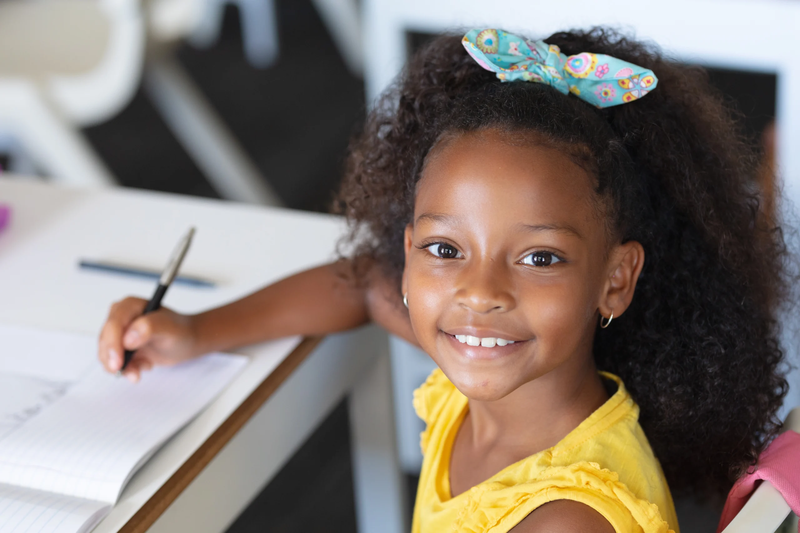 A smiling little girl sitting at a desk, writing with a pencil.