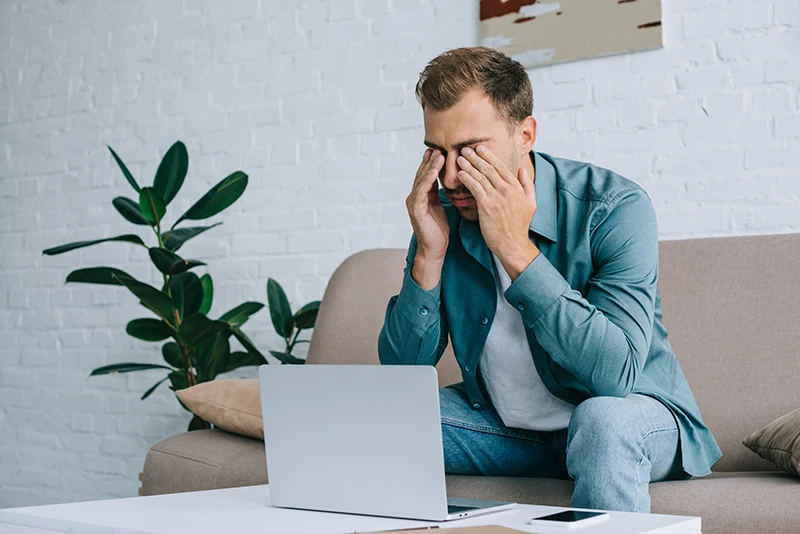 a young man rubbing his eyes while working on a laptop at home